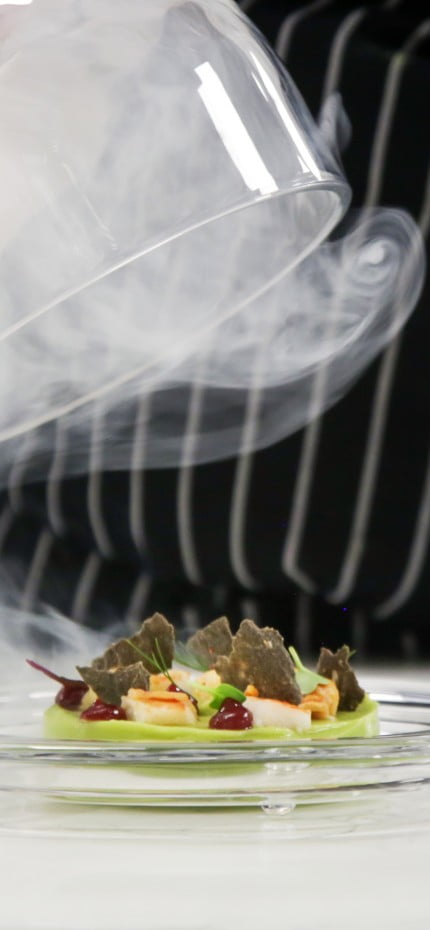 Chef's hand lifts up glass cloche from a plate with hot food and moving smoke at the restaurant.
