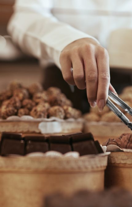Caramelos De Chocolate En Tienda De Confitería. Primer Plano De Mano Femenina Con Fórceps Escogiendo Caramelos. Alta resolución