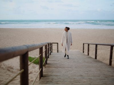 Mujer solitaria caminando por la playa camino de madera. Serena afroamericana disfrutando del tiempo a orillas del océano. Modelo de pelo negro paseando en un día nublado. Muchacha hermosa sonriente que toma la rotura que va el lugar pacífico solo.