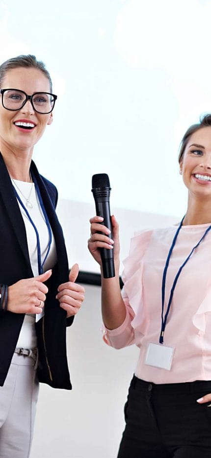 Dos mujeres jóvenes mantienen una conversación en público, una de ellas sostiene un micrófono.