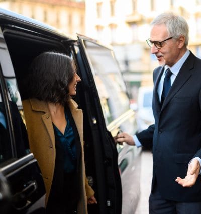 Chauffeur opening the door of his vehicle while welcoming his passenger to her destination.