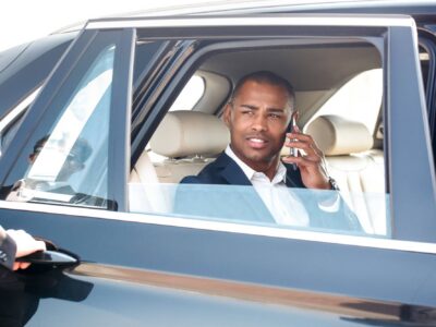 Security guard opening door young businessman sitting in the car talking with friend on smartphone looking aside smiling happy