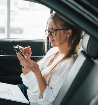 Mujer de negocios consultando un dispositivo móvil desde el asiento trasero de un vehículo de alta gama.