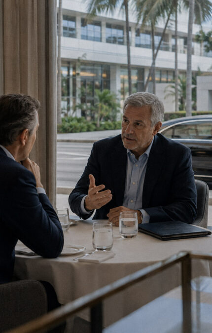 Two businessmen discussing business at a table, probably at a restaurant, while an executive vehicle is visible outside from the glass window.